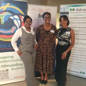 Three professional women smiling and posing in front of business organization banners at a networking event.