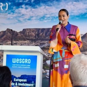 Woman in colorful dress speaking into a microphone at a Wesgro European Trade & Investment event with mountains and blue sky backdrop.